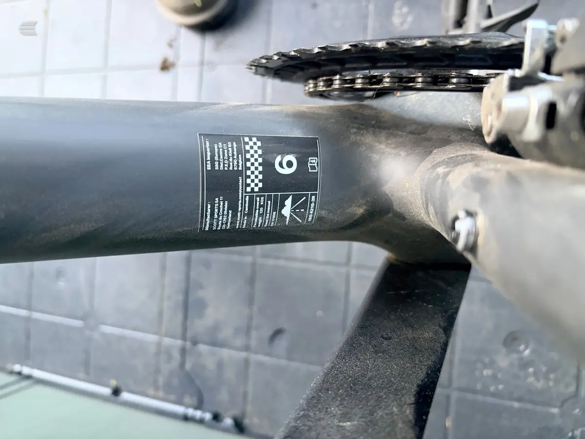 Close-up of a bicycle seatpost with a black label and checkered branding detail, mounted on a dark frame against a tiled background.