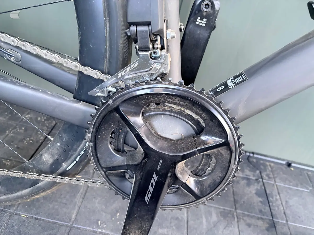 Shimano crankset and chainring on a grey road bike, drive side close-up, indoors against a pale green wall.