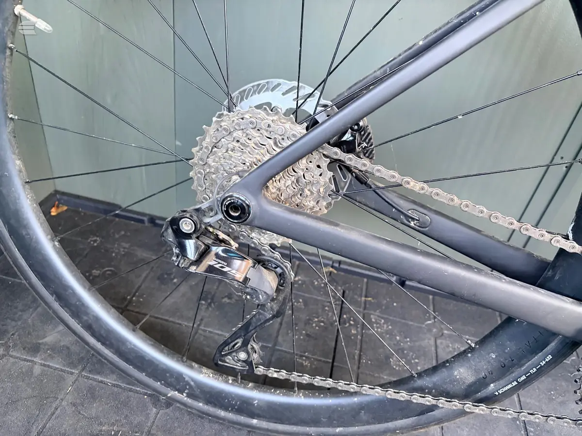 Close-up of bicycle rear derailleur, cassette and chain on silver frame, drive side, indoors against dark floor.