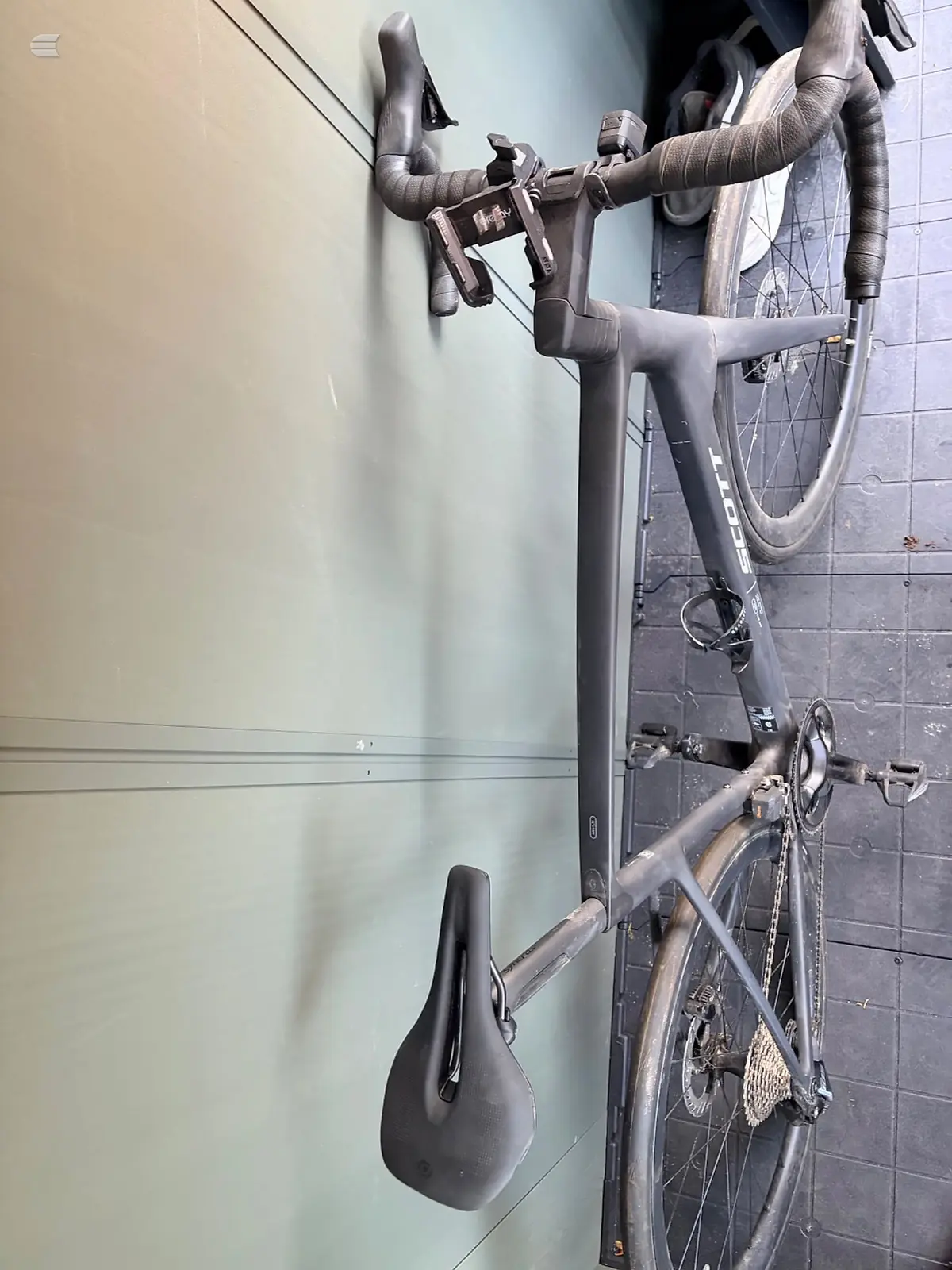 Grey road bike with drop bars and wrapped handlebar tape, mounted on wall against light and dark tiled background, non-drive side view.