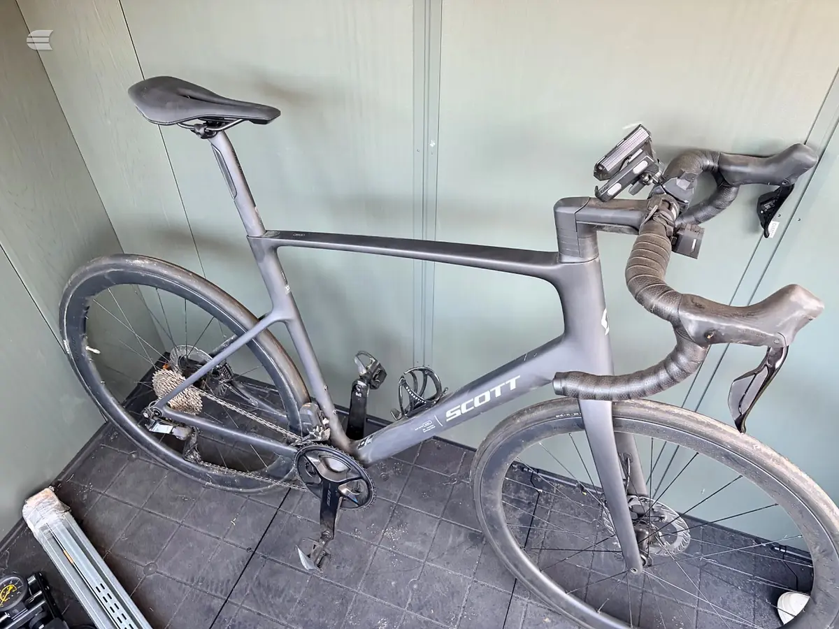 Grey Scott road bike with drop bars, non-drive side, leaning against white wall in indoor setting.
