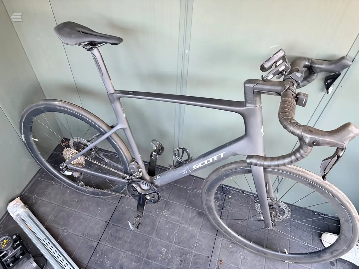 Scott road bike, grey/purple, non-drive side, leaning against white wall in garage setting with drop bars and saddle visible.