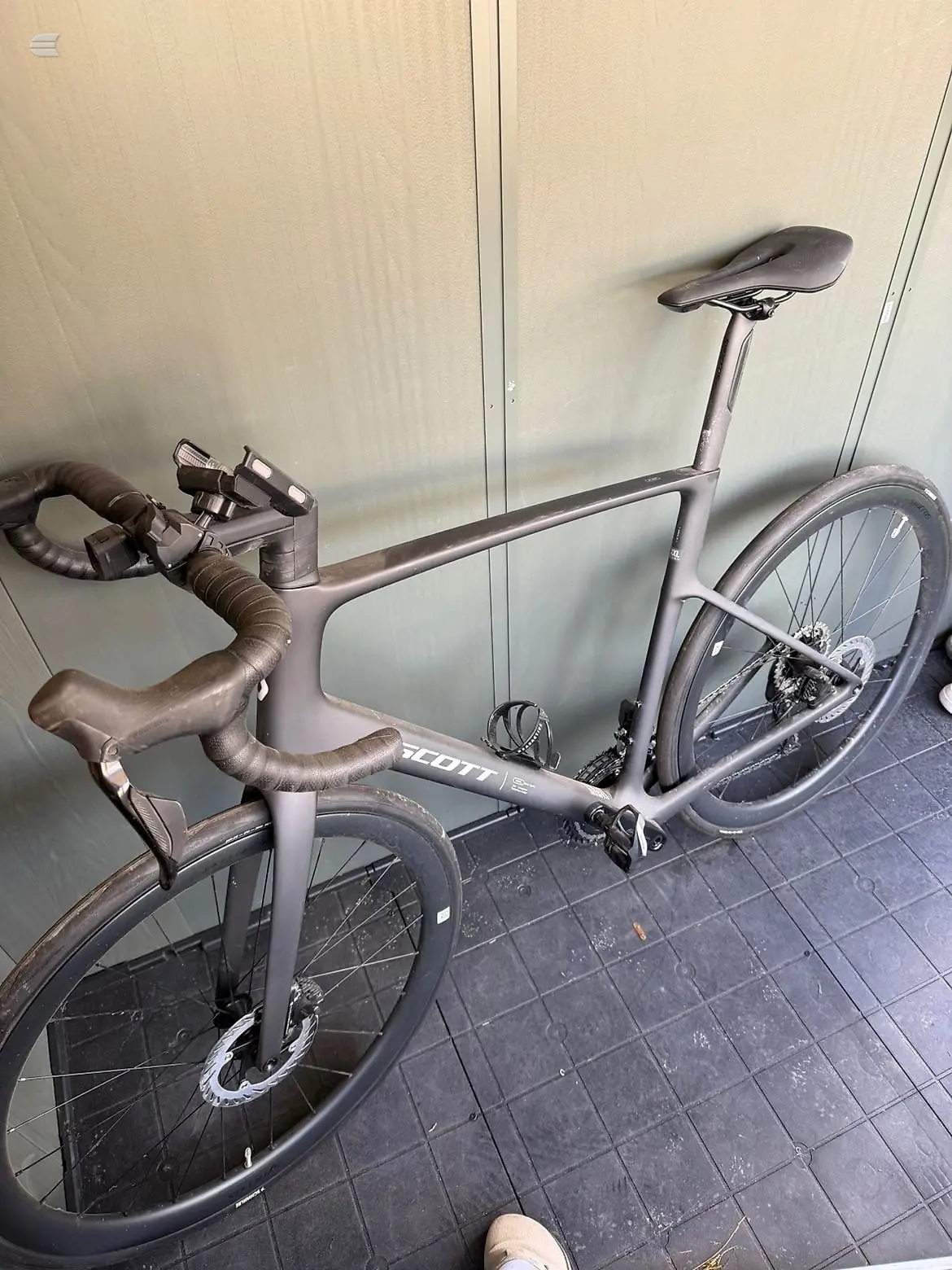 Grey Scott road bike with drop bars, drive side, leaning against a white corrugated wall indoors.
