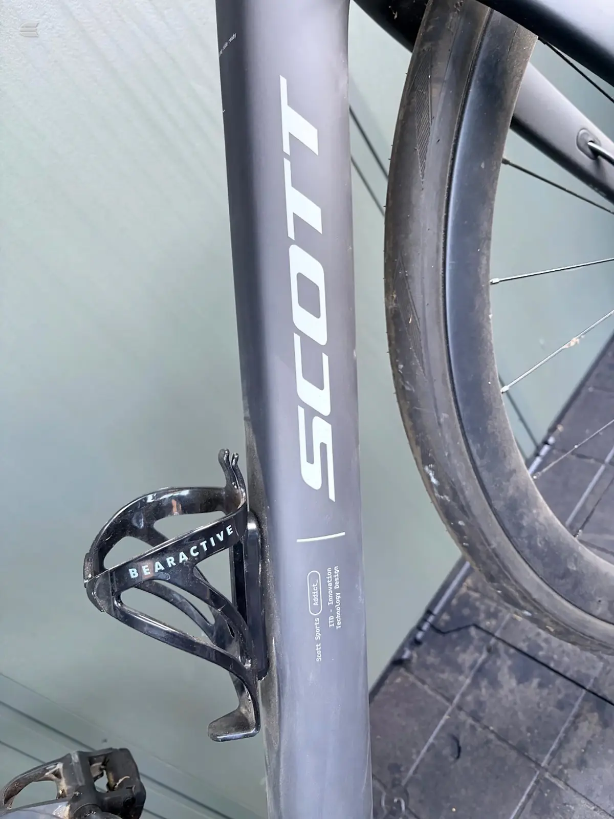 Grey Scott road bike frame, non-drive side, close-up of seat tube with Scott branding and Bearactive bottle cage mounted. Indoor setting against white wall.