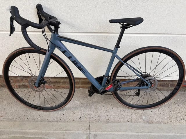 Blue gravel bike with drop bars and disc brakes, drive side, against a white rendered wall and brick base.