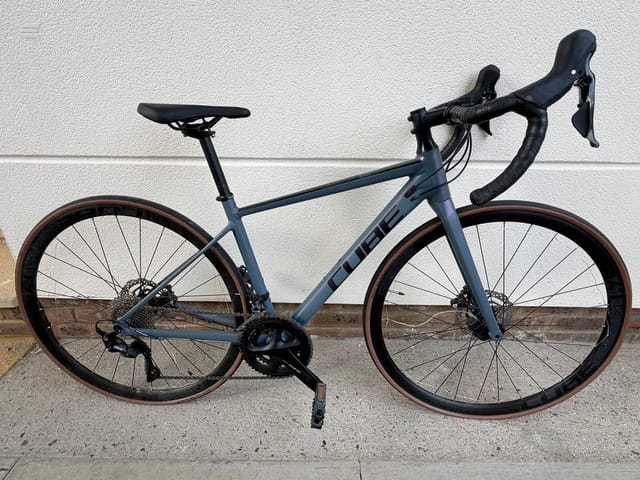 Blue-grey gravel bike on kickstand, drive side, against white textured wall and concrete base.