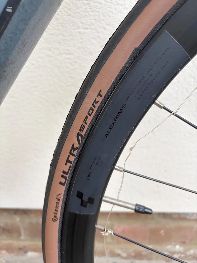 Close-up of two bicycle wheels side by side, non-drive side view. Brown Continental Ultra Sport tyre on left, black Alexrimes rim on right, against white