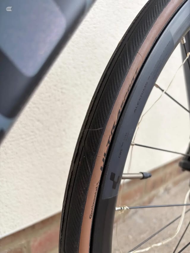Close-up of bicycle wheel rim and tyre, angled view against white wall, showing spoke detail and brake components.