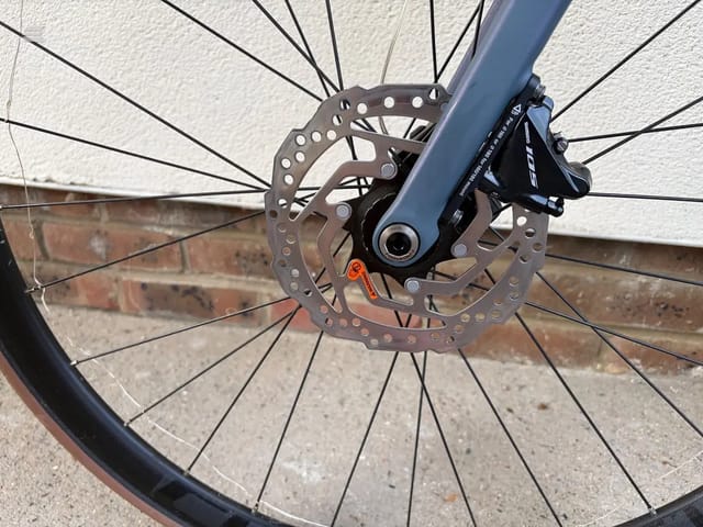 Close-up of front disc brake wheel on blue-grey gravel bike, drive side, against brick wall and pavement.