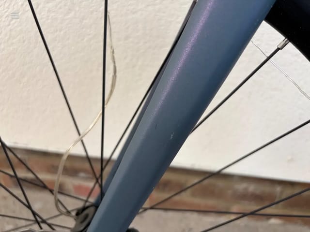 Close-up of blue bicycle frame tubing and spokes against white wall background, detail shot of head tube and fork area.