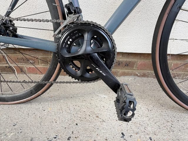 Close-up of black triple crankset and pedal on a road bike, drive side, against a brick wall and pavement.