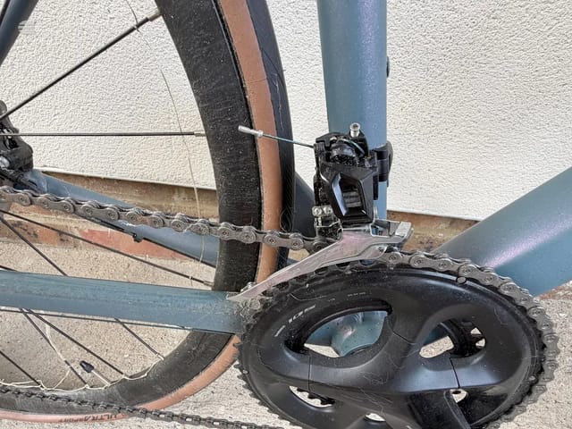 Close-up of bicycle crankset and front derailleur, drive side, showing chain and triple chainring against textured wall background.
