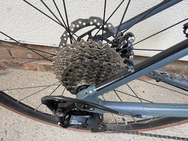 Shimano rear derailleur and cassette on blue road bike, drive side close-up against concrete wall.