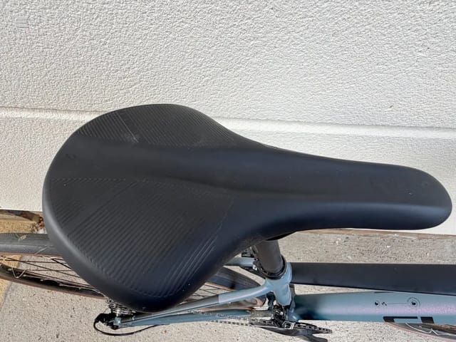 Black saddle on a bicycle frame, close-up side view against a textured white wall, showing seat post and chain stay.