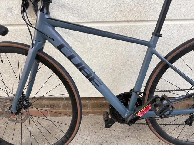 Blue-grey gravel bike with brown tyres, drive side, leaning against a white brick wall and brown brick base.