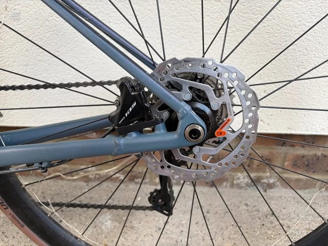 Close-up of disc brake system on blue gravel bike, drive side, showing hydraulic caliper and rotor against concrete background.