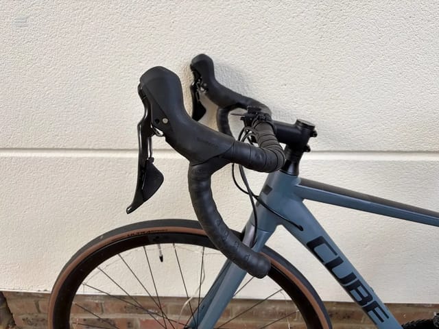Blue-grey gravel bike with drop bars and brown tyres, drive side, against a textured white wall.