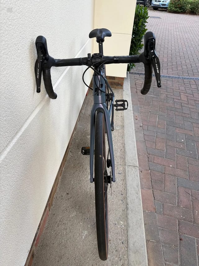 Black drop-bar road bike, front-on view, parked against a white and cream rendered wall on a brick paved driveway.