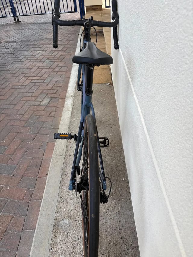 Blue road bike with drop bars, viewed from above along its length, parked in a narrow alley between a white wall and brick paving, drive side visible.