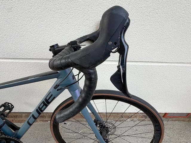 Blue-grey Cube road bike with drop bars, drive side, against a white textured wall. Shimano components visible.