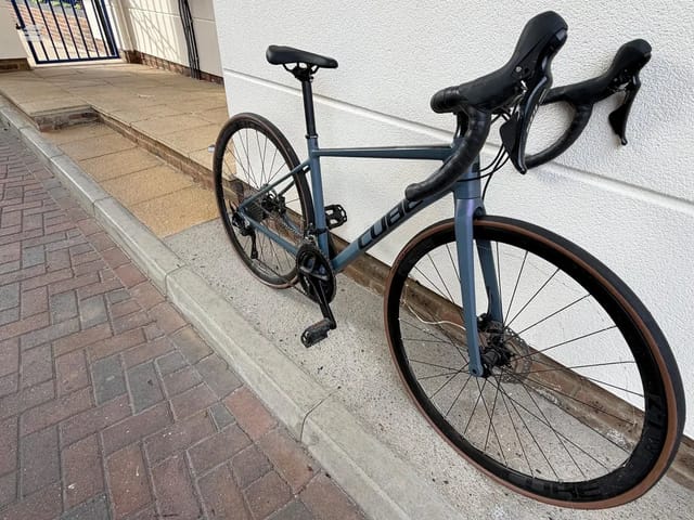Blue-grey Cube gravel bike with drop bars, drive side, leaning against white wall on residential driveway.