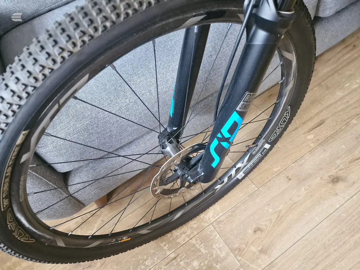 Close-up of front wheel and fork on a gravel bike with disc brakes, indoors on wooden floor, non-drive side view.