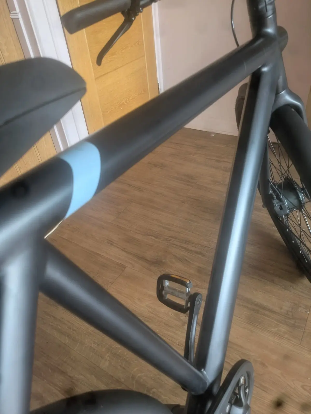 Black and blue stationary exercise bike indoors on wooden floor, non-drive side, against wooden door and beige wall.