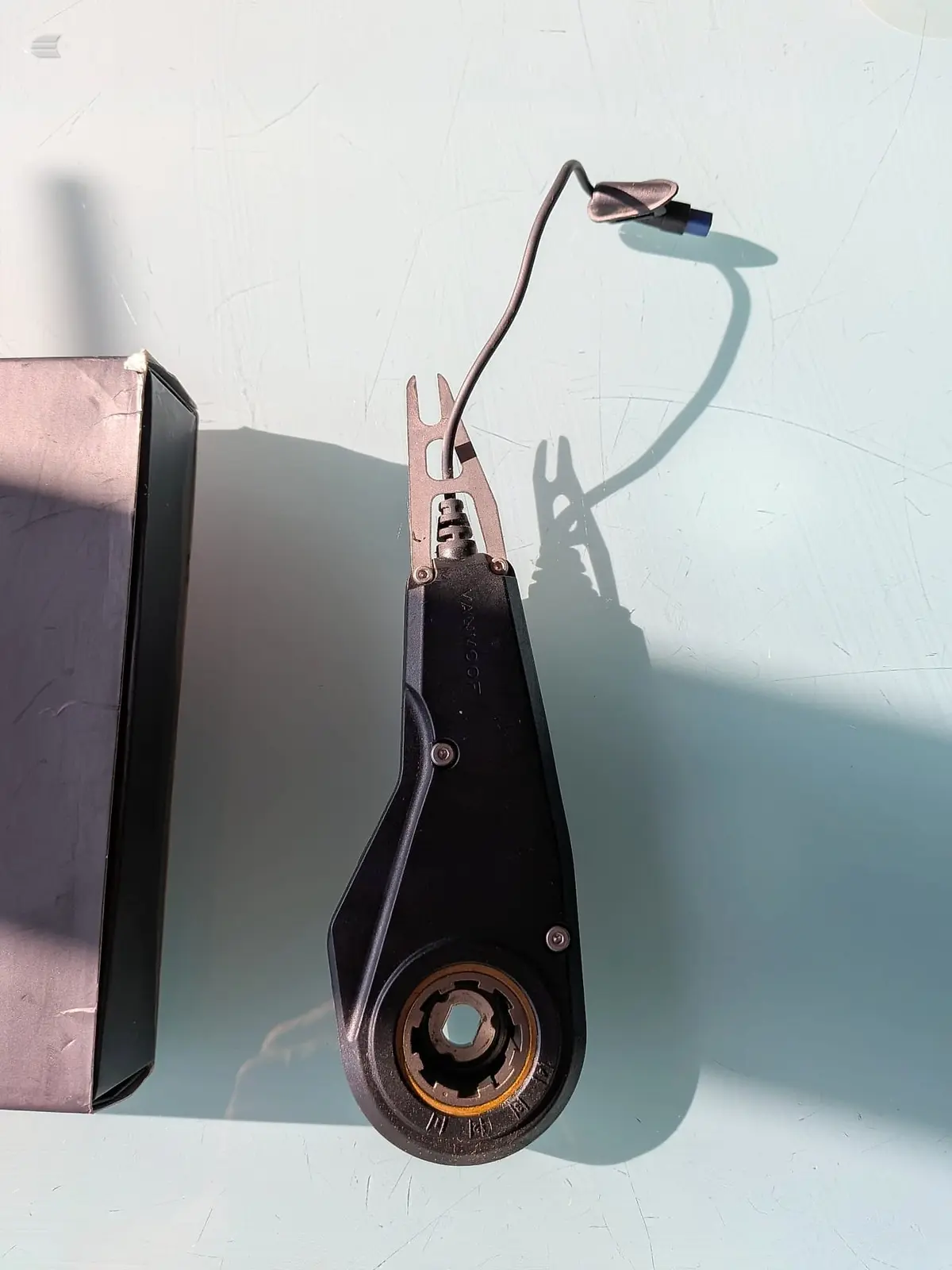 Vintage bicycle brake caliper and cable assembly, copper and black metal, mounted vertically against white textured wall with shadow detail visible.