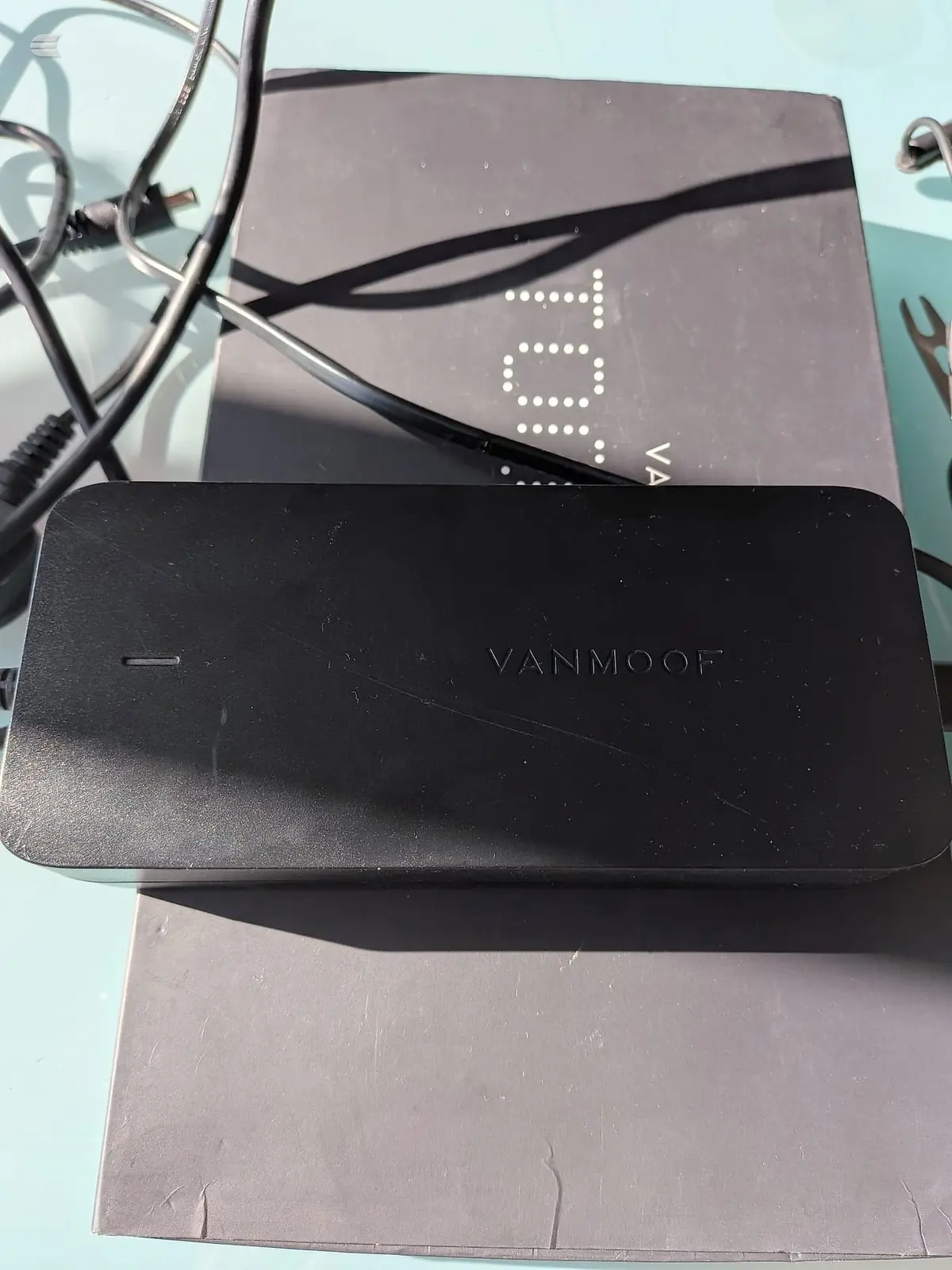 Two black electronic devices stacked on a light blue surface with power cables, branded VANMOOF, against a cardboard box.