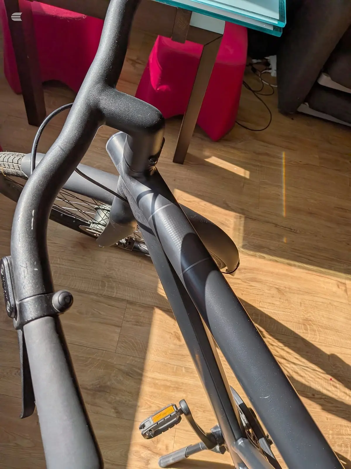 Grey road bike on wooden floor, drive side, angled toward camera. Red cushioned furniture and teal shelving visible in background.