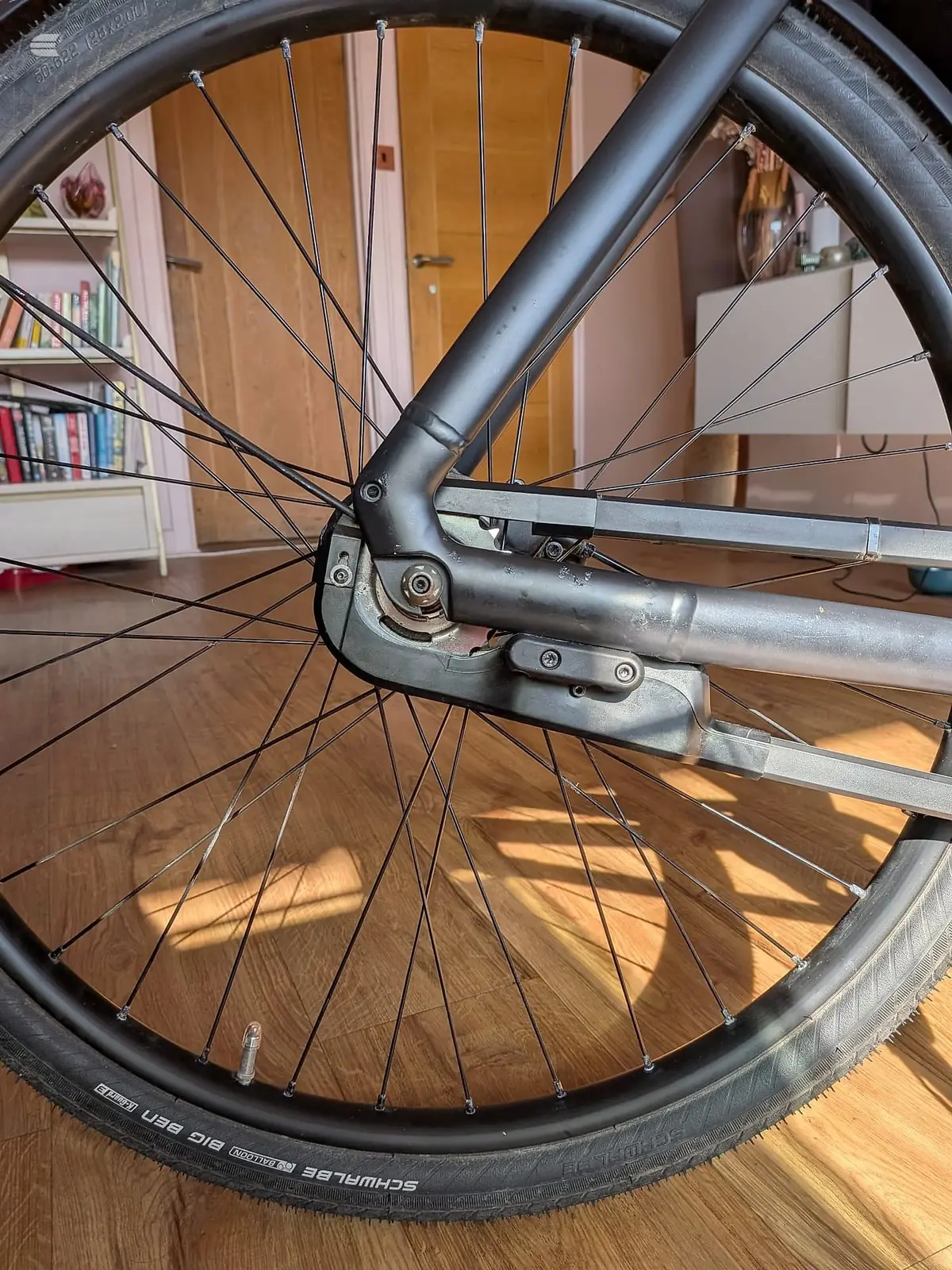 Close-up of bicycle wheel with spokes and brake caliper, shot indoors on wooden floor with bookshelf and wooden door visible in background.