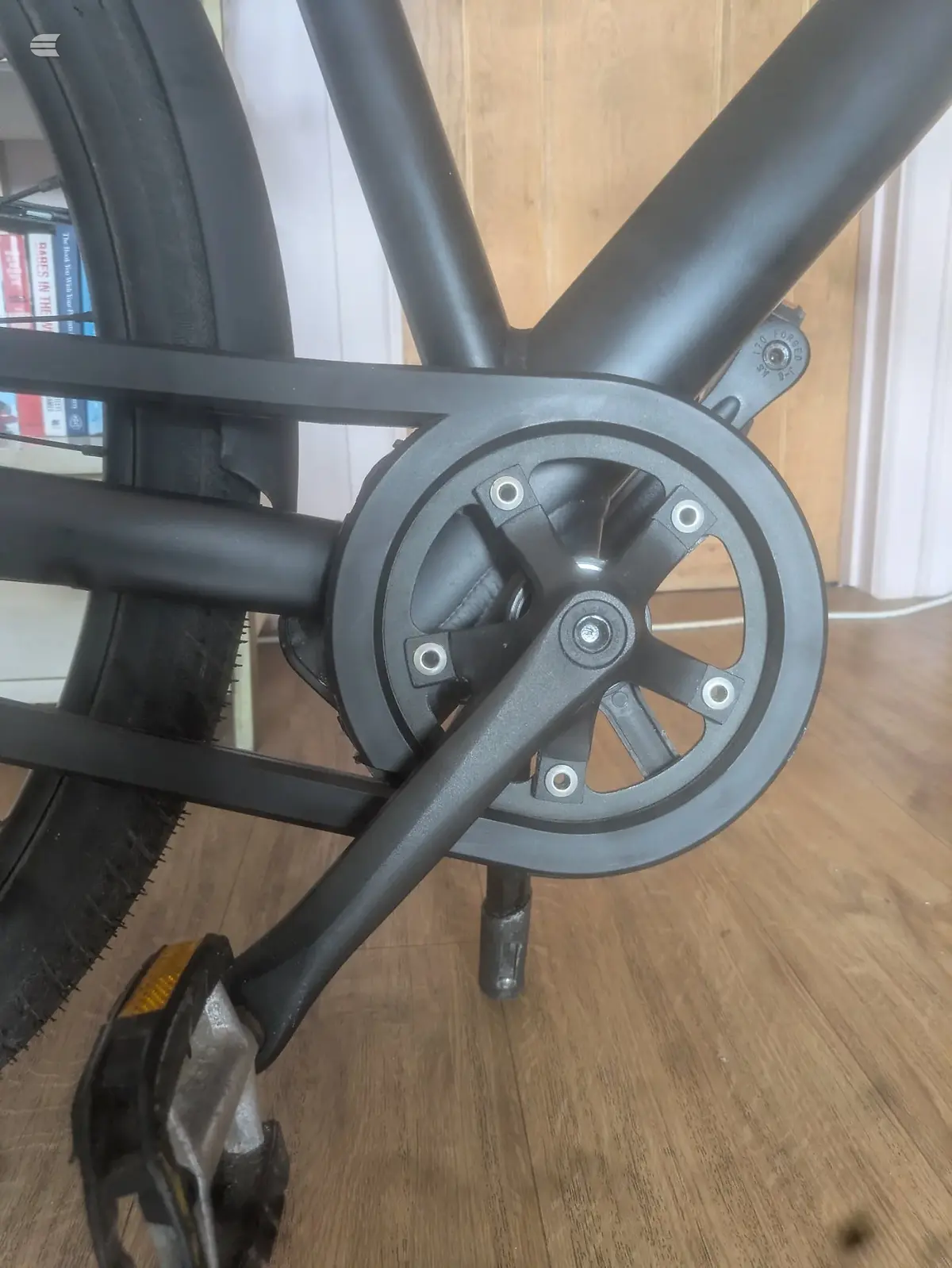 Black stationary bike crank and chainring close-up, indoor wooden floor setting, drive side angle.