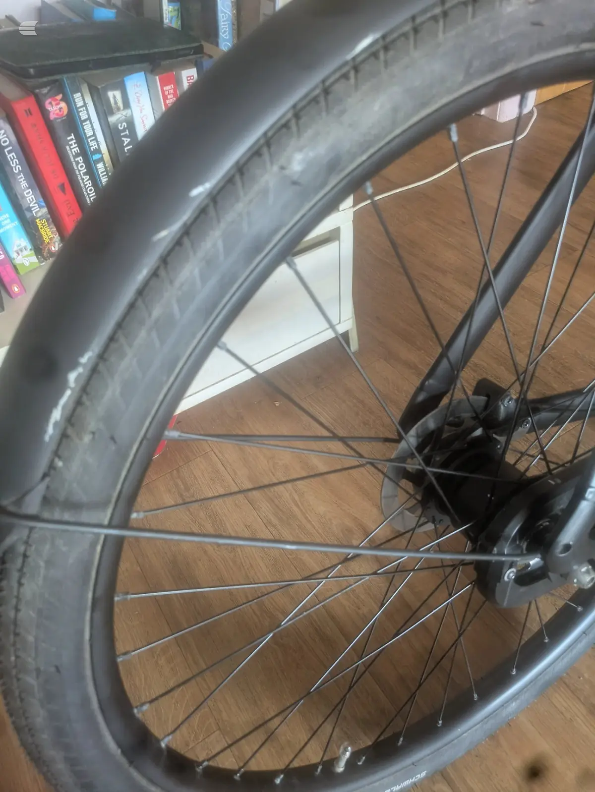 Close-up of a bicycle wheel with dark tyre and spokes, indoors on wooden floor with bookshelves visible in background.