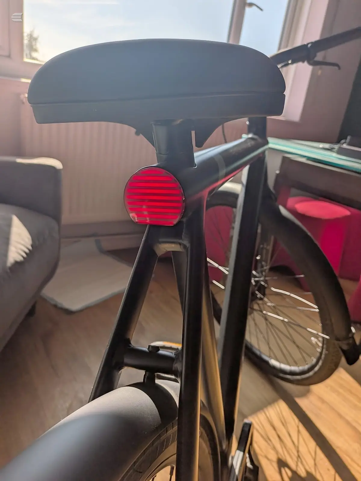 Black saddle and red rear light on a black-framed bike, indoor setting with wooden floor and pink wall, rear three-quarter view.