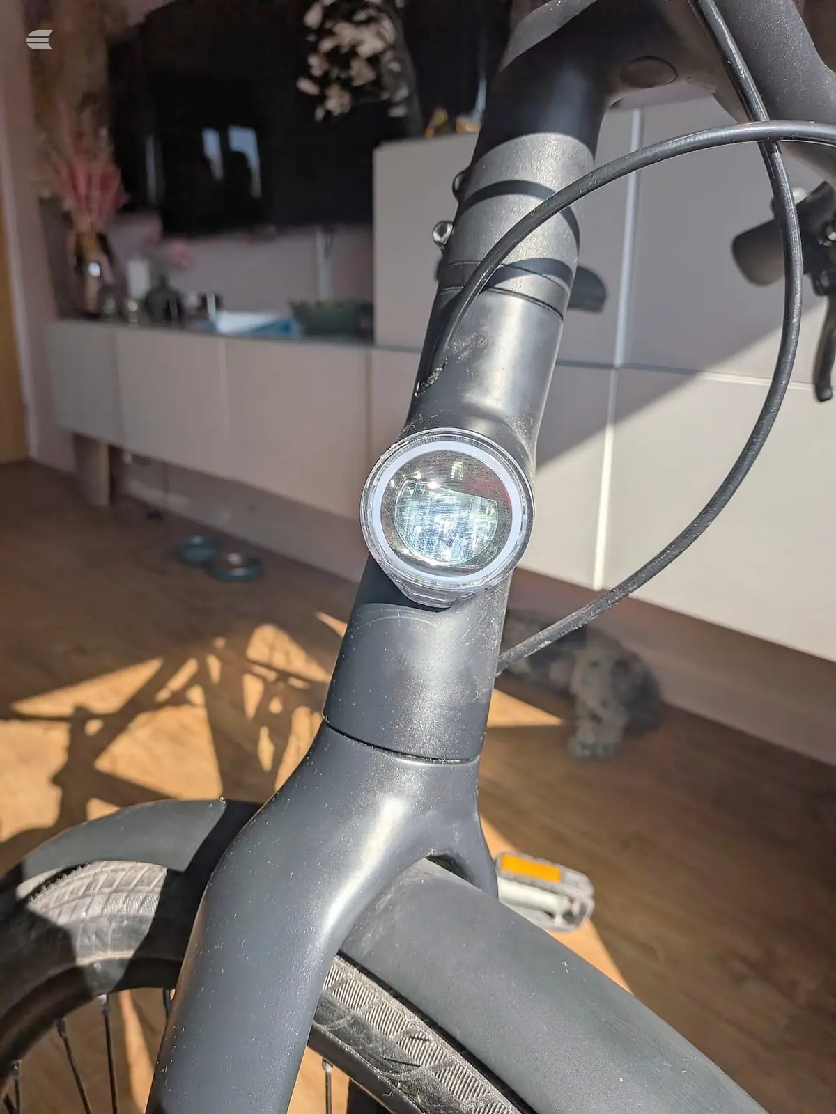 Close-up of a bicycle headlight mounted on the stem of a dark-coloured bike, indoors on a wooden floor with white furniture in the background.