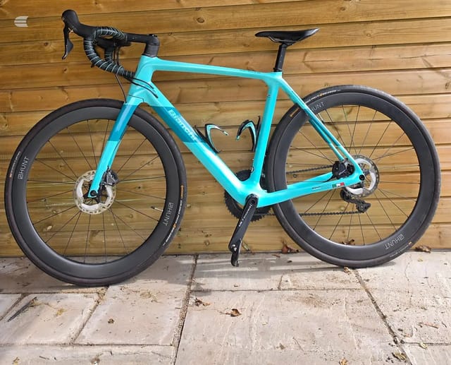 Bright turquoise gravel bike with disc brakes and deep-section black wheels, displayed side-on against a wooden fence on a paved surface.