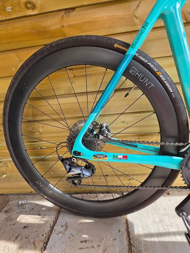 Turquoise gravel bike with deep-section dark rims, displayed drive-side on wooden decking. Front wheel in sharp focus filling most of frame, rear wheel and