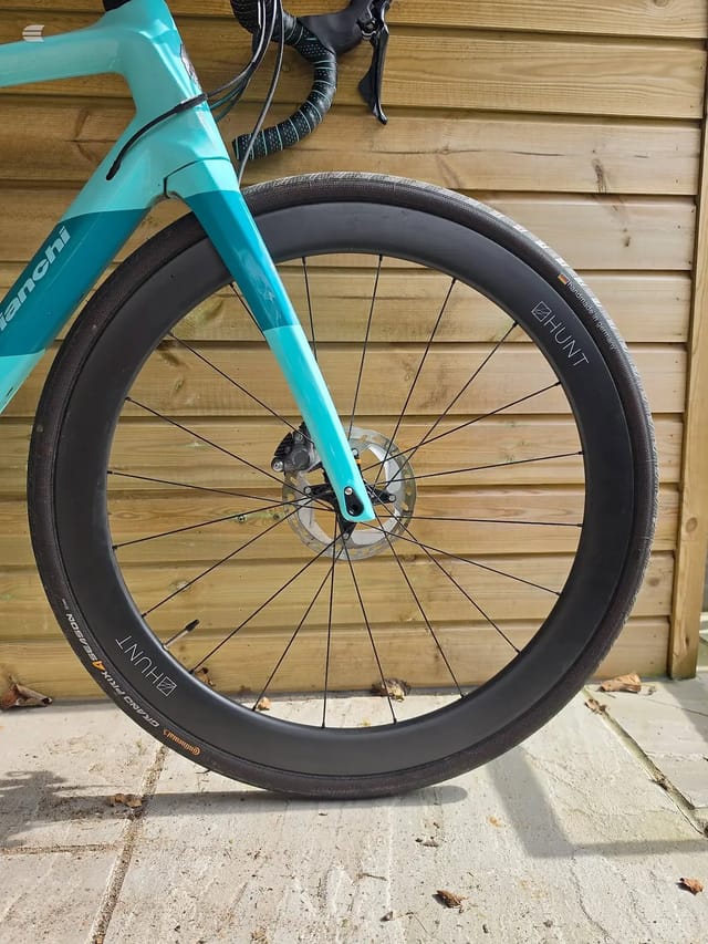 Turquoise gravel bike front wheel with deep-section black rim, disc brake, and drop bars visible, on concrete against wooden fence, non-drive side.
