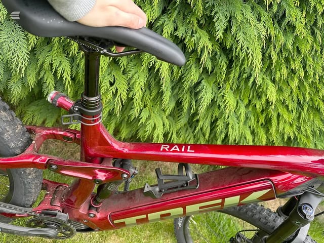 Red Rail hardtail mountain bike, three-quarter view from above, drive side, against bright green conifer foliage. Hand on saddle.