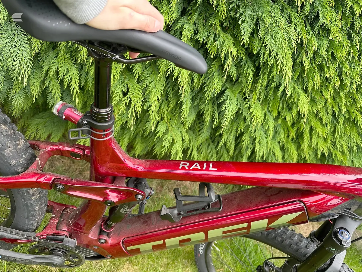 Red Rail hardtail mountain bike, three-quarter view from above, drive side, against bright green conifer foliage. Hand on saddle.
