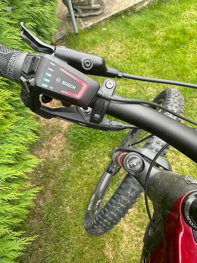 Bosch e-bike motor and display unit mounted on handlebars, close-up view on grass background with bike frame and front wheel visible.