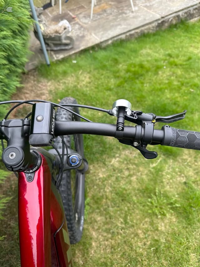 Close-up of red electric bike handlebars and cockpit, non-drive side, showing brake lever, bell, and display unit against a garden background.