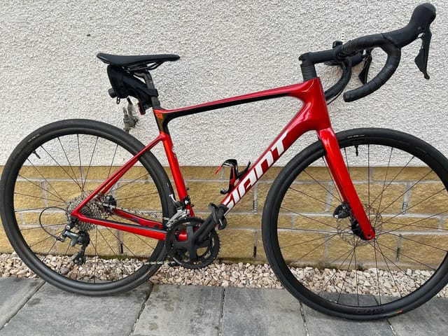 Red Sram road bike with black drop bars and saddle, drive side, leaning against a textured grey and tan wall on gravel.