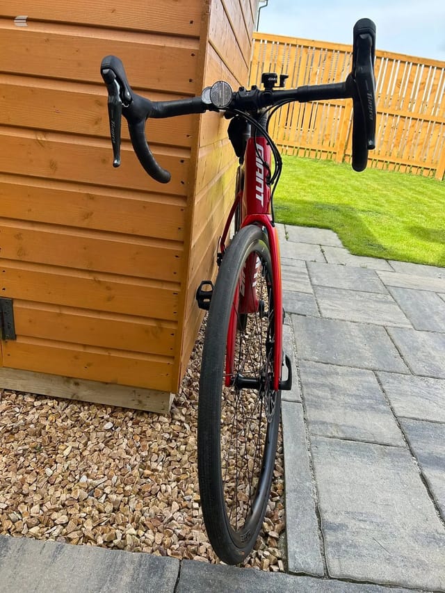Red drop-bar road bike leaning against wooden shed, drive side, paved garden setting with yellow fence visible behind.