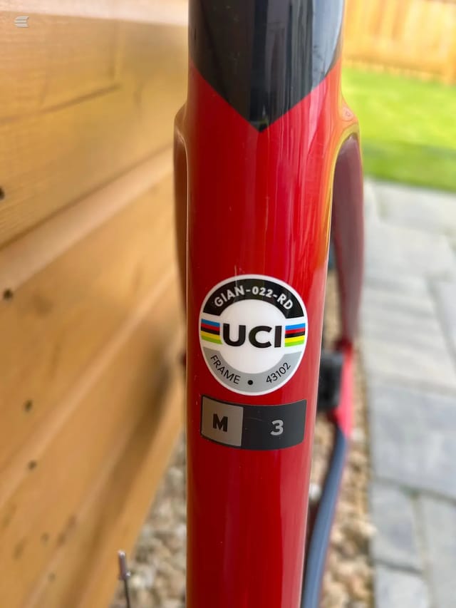 Close-up of red Giant road bike frame tube with UCI sticker, model marking M3, against wooden garage door and garden background.