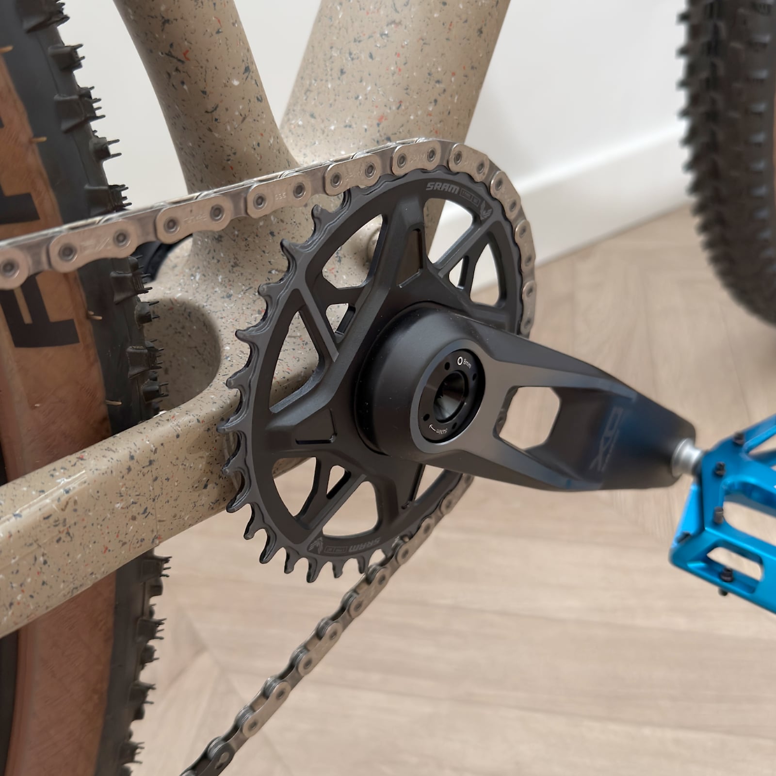 Close-up of SRAM crankset and chainring on tan gravel bike frame, drive side, wooden floor background.