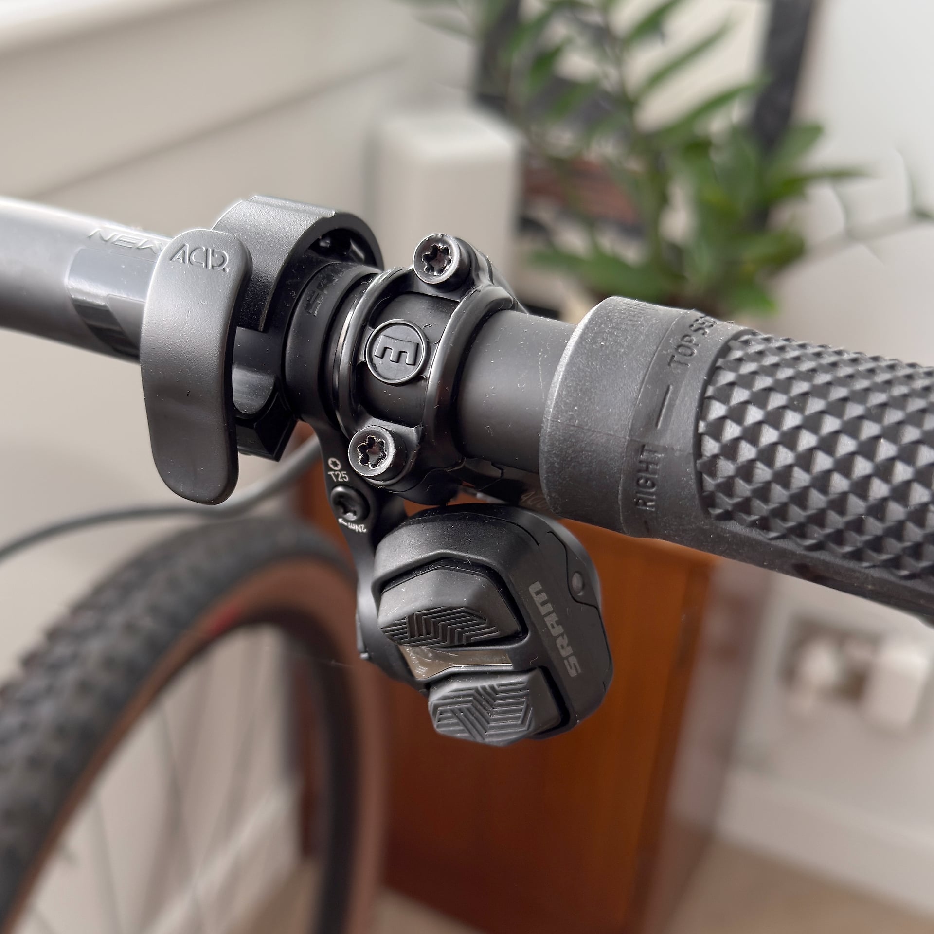 Close-up of bicycle handlebar stem and brake lever assembly, black components on textured grip, indoor setting with blurred plant and furniture background.