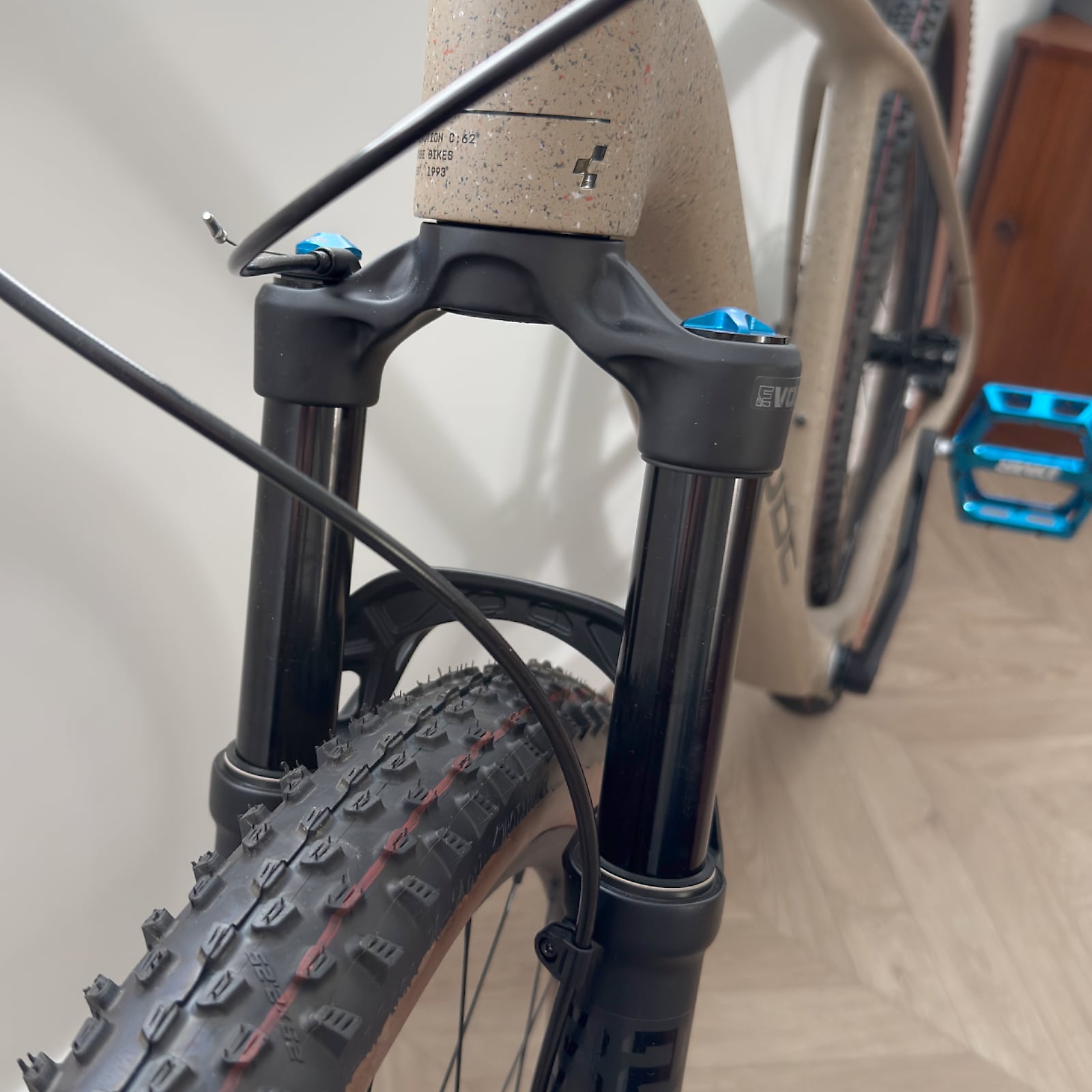 Black suspension fork with blue damper adjusters and cork grip tape on tan gravel bike, close-up detail shot indoors.