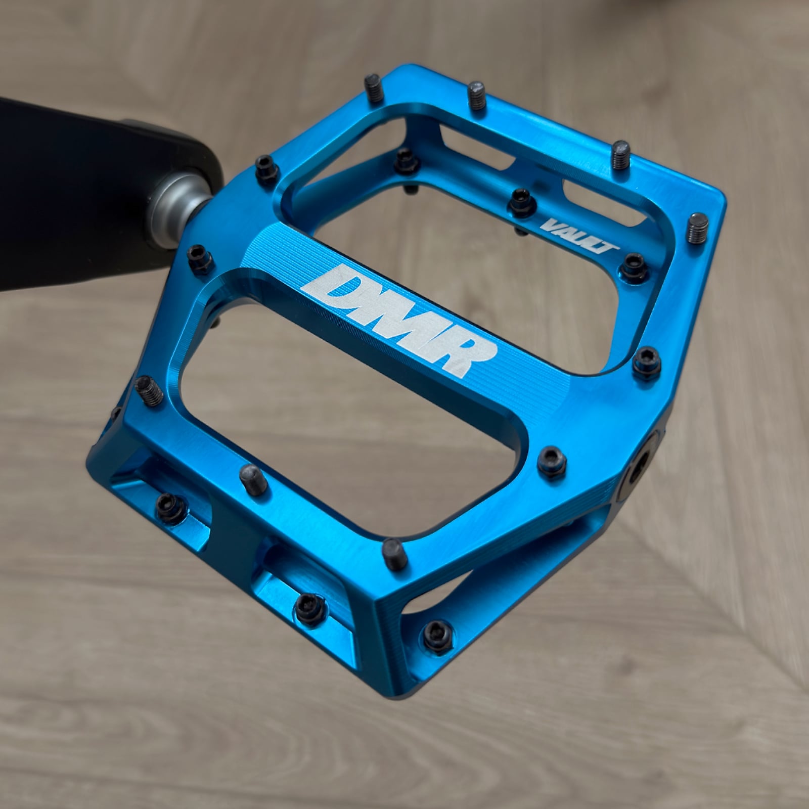 Blue DMR platform pedal with textured grip surface and black cleats, shown from above against a neutral beige background.