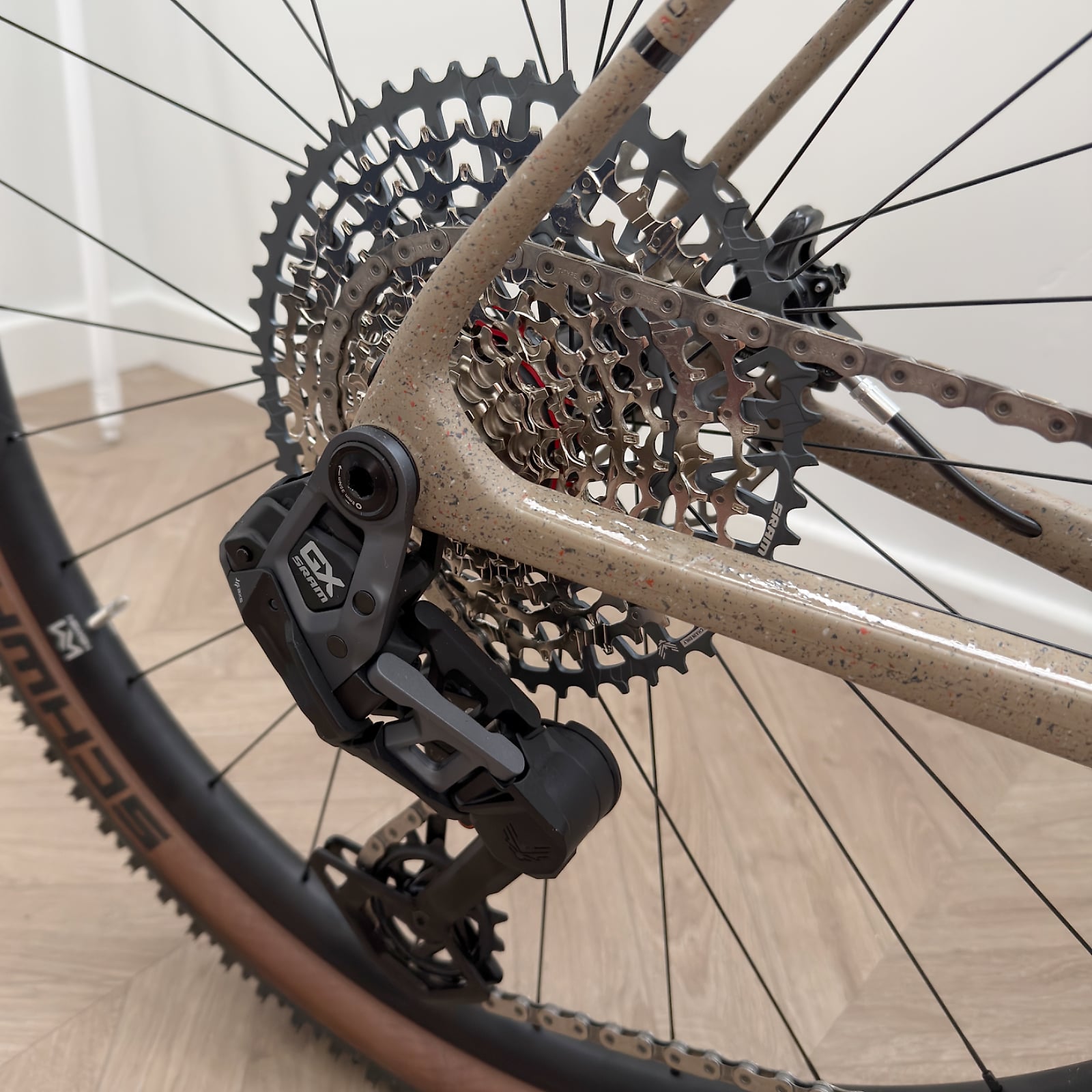 Close-up of tan gravel bike rear drivetrain, drive side, showing cassette and derailleur indoors on wooden floor.